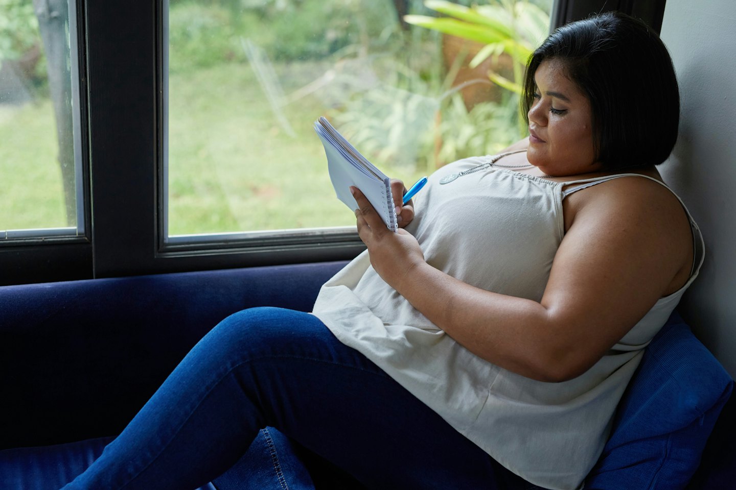 Woman writes in notebook on a blue sofa by a window.
